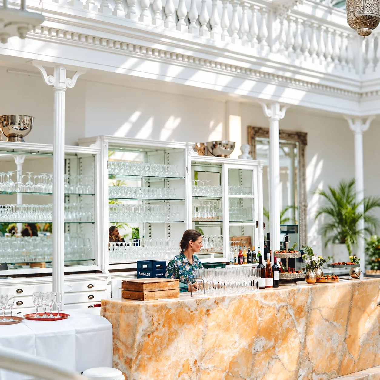 Die Bar mit Marmortresen und Gläservitrinen unter der weißen Galerie, im Sonnenlicht.